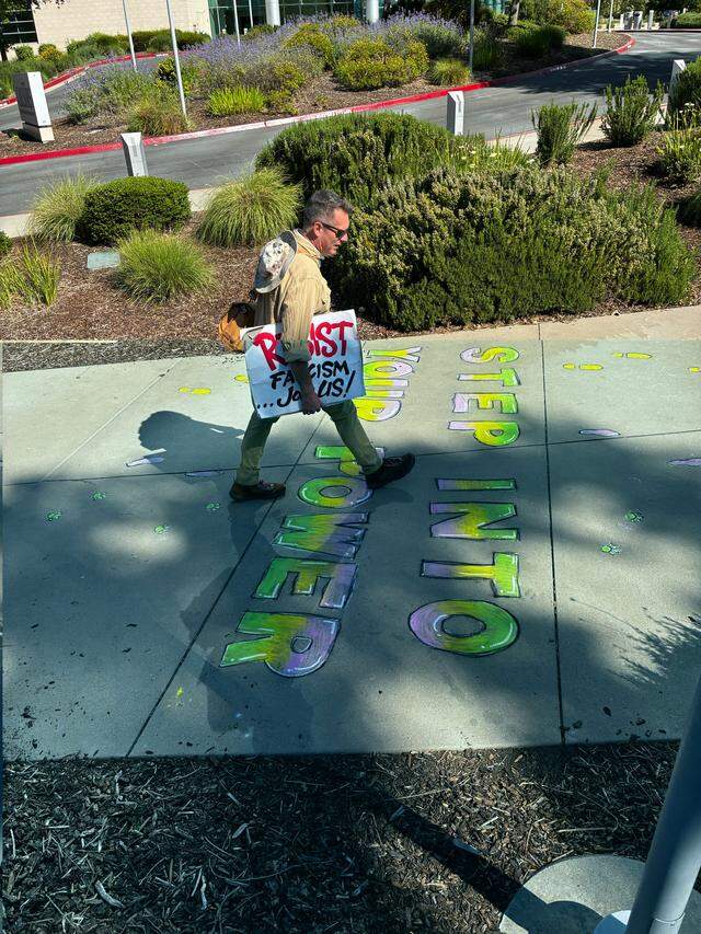 A protester arriving at West Sacramento’s River Walk Park walks over sidewalk chalk that reads “Step into your power” before a “No Kings” rally Saturday morning.