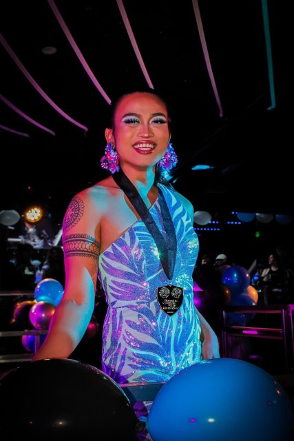 Person in a sparkling blue dress with bold makeup and earrings, wearing a medal, stands in a decorated venue with black and blue balloons.