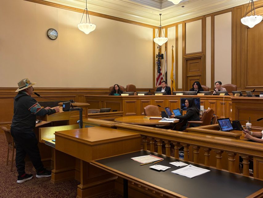 A person speaks at a podium in a wood-paneled courtroom or government chamber, addressing a panel of officials seated behind desks with microphones and computers.