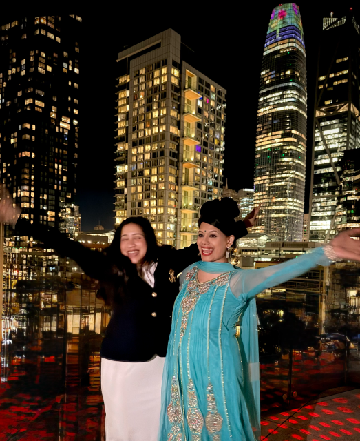 Two women pose with arms outstretched on a rooftop at night, with tall, illuminated city buildings in the background.