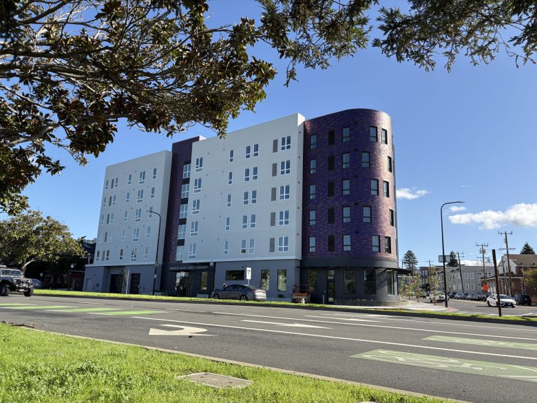 A six-story apartment building is seen from the opposite side of a broad street. Part of its facade is a rounded corner of purple bricks.
