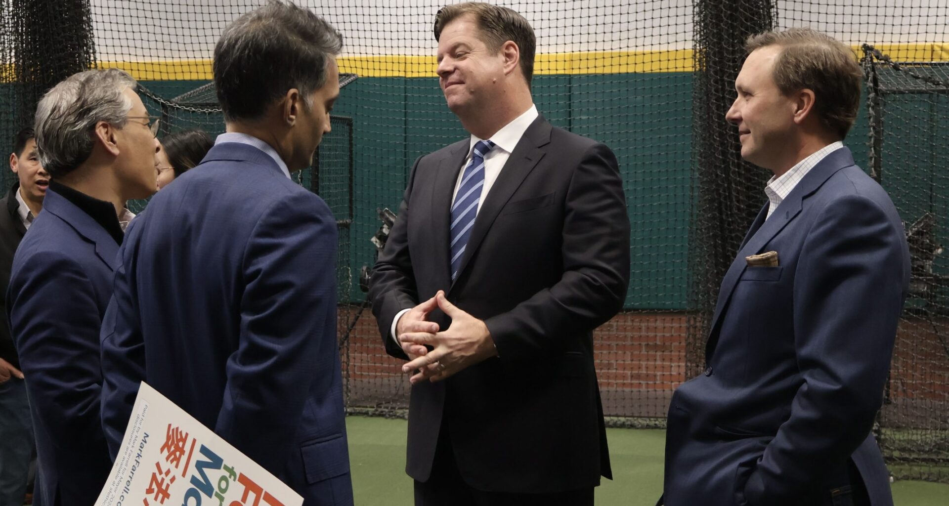 Four men in business attire are conversing in an indoor setting, with one holding a sign. A netted wall and green flooring are visible in the background.