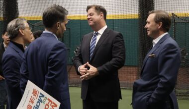 Four men in business attire are conversing in an indoor setting, with one holding a sign. A netted wall and green flooring are visible in the background.