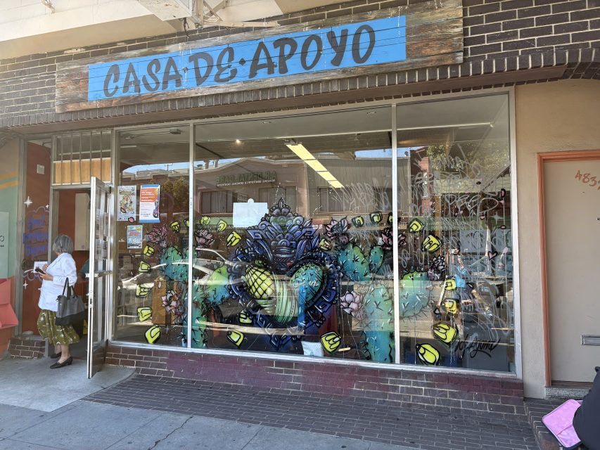 Storefront with a "Casa de Apoyo" sign, large window featuring colorful mural art, and people entering and exiting the building.