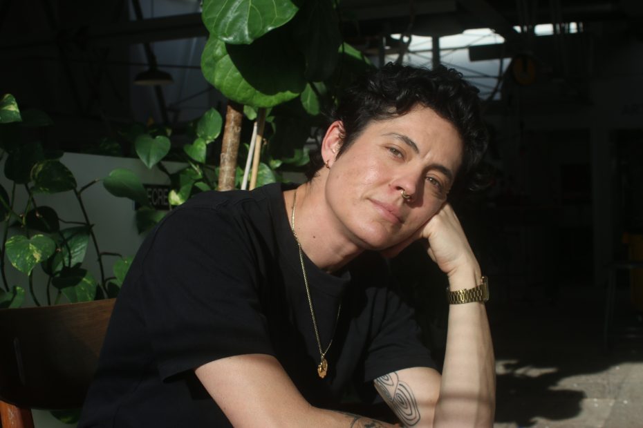 Person with short dark hair, wearing a black shirt, gold necklace, and watch, sits indoors by green plants, resting their head on their hand and looking at the camera.