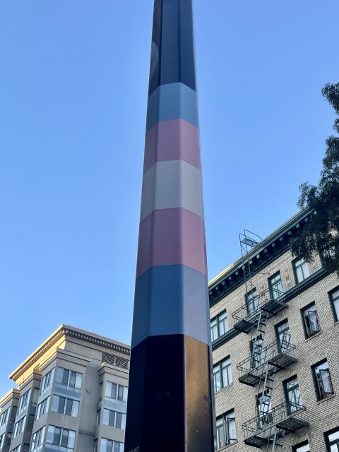 A tall pole painted with horizontal stripes of black, blue, pink, white, and brown. It is set against a clear blue sky and adjacent to multi-story buildings.