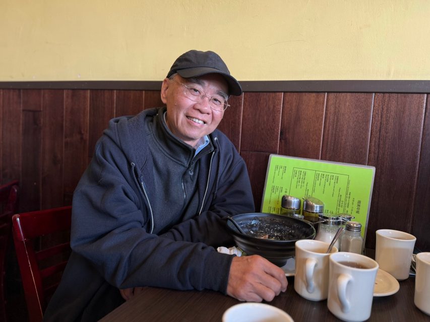 A man wearing glasses and a cap sits at a restaurant table with a bowl of food, four cups, condiments, and a green menu.