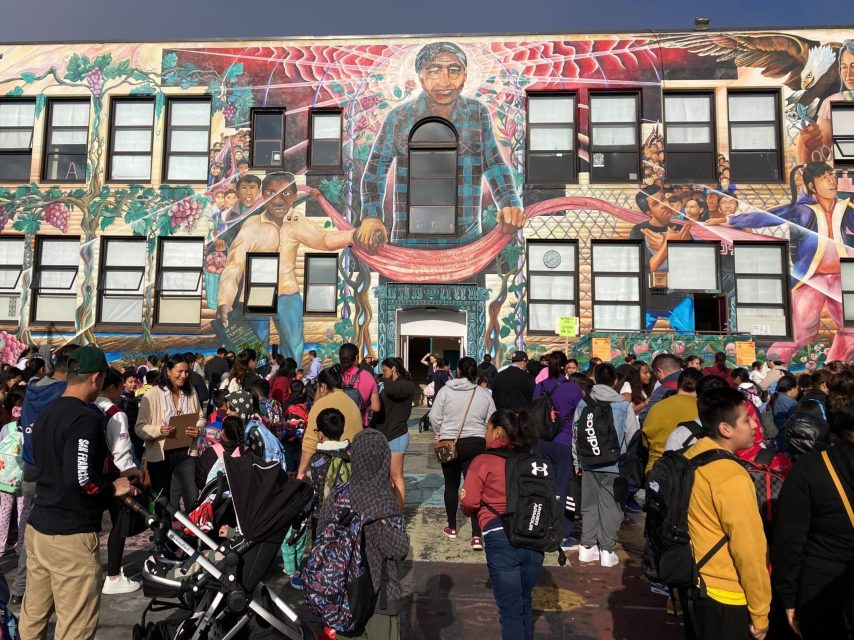 A large group of people, including many children, gather in front of a mural-covered building on a sunny day. The mural depicts various figures and vibrant scenes.