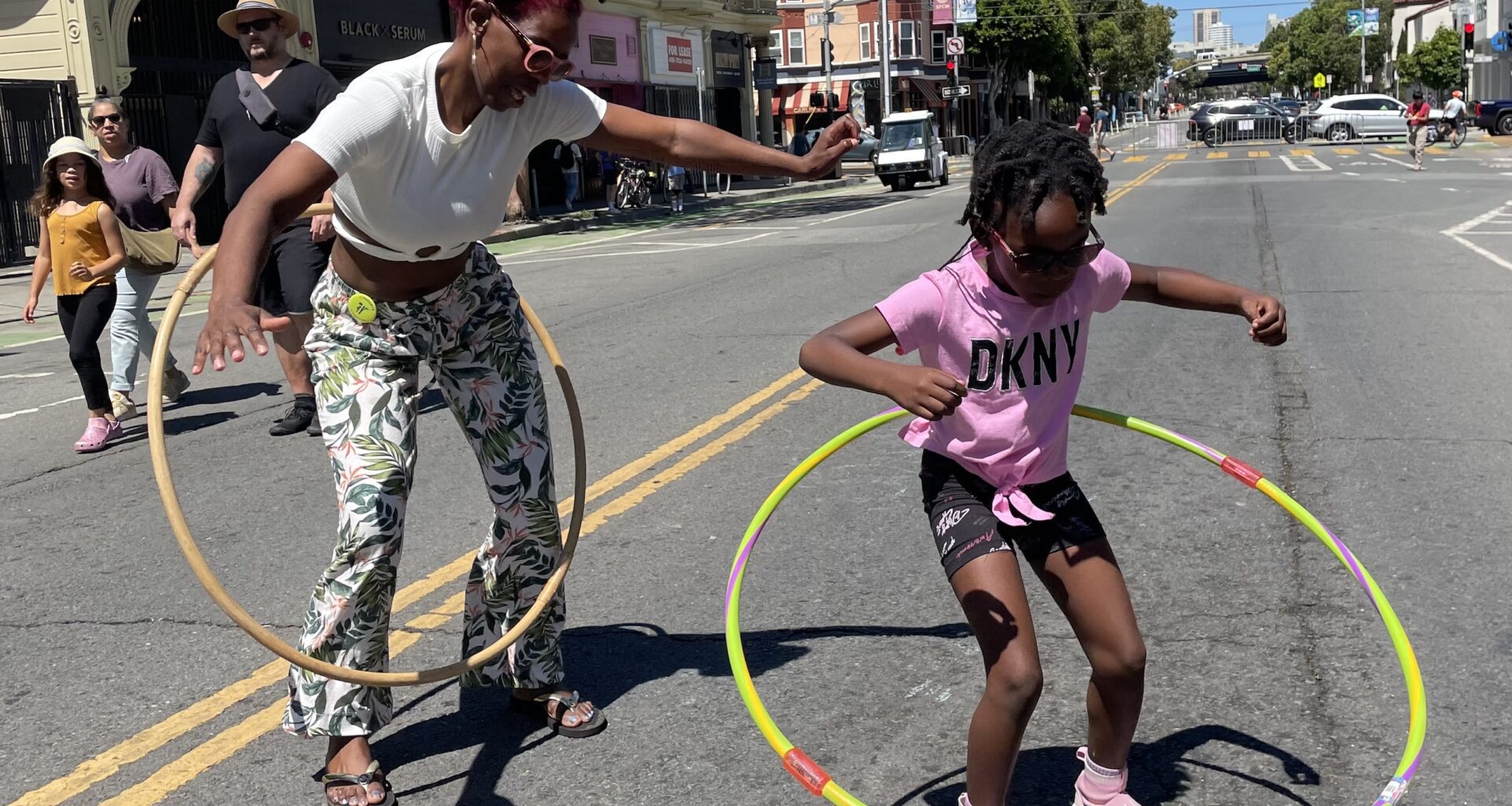 Two people hula hoop on a sunny street with buildings and pedestrians in the background. One is an adult wearing patterned pants, and the other is a child in a pink shirt and shorts.