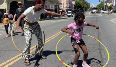 Two people hula hoop on a sunny street with buildings and pedestrians in the background. One is an adult wearing patterned pants, and the other is a child in a pink shirt and shorts.