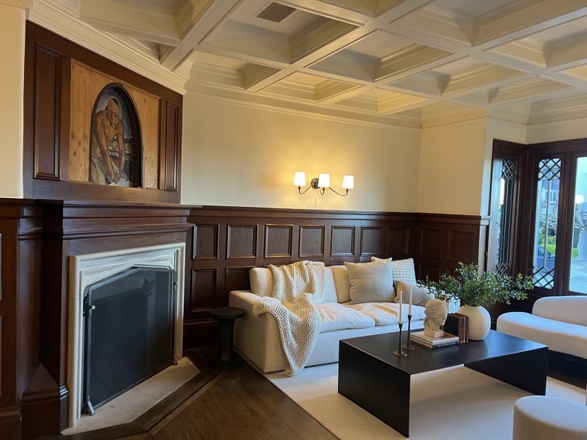 A living room with a coffered ceiling, wood paneling, fireplace, light sofa, black coffee table, and wall-mounted artwork above the mantel.