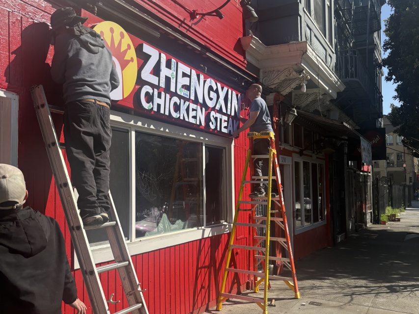 Two workers on ladders install a "Zhengxin Chicken Steak" sign on the exterior of a red building along a sidewalk in daylight.