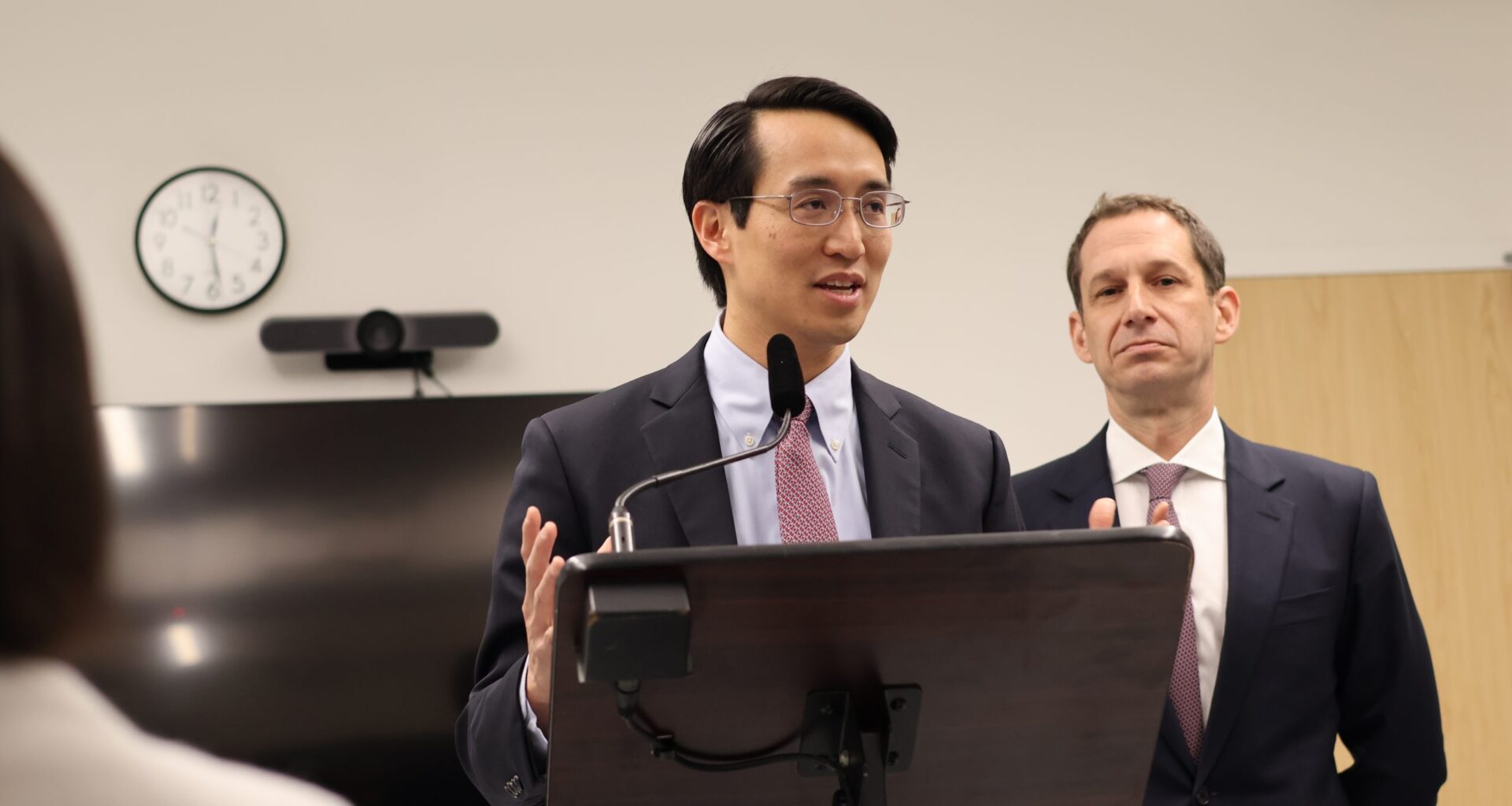 A person speaking at a podium with a microphone, wearing a suit and tie. Another person stands behind, also in a suit. A wall clock and TV are in the background.