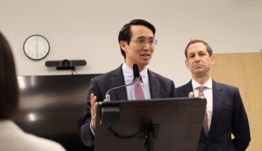 A person speaking at a podium with a microphone, wearing a suit and tie. Another person stands behind, also in a suit. A wall clock and TV are in the background.