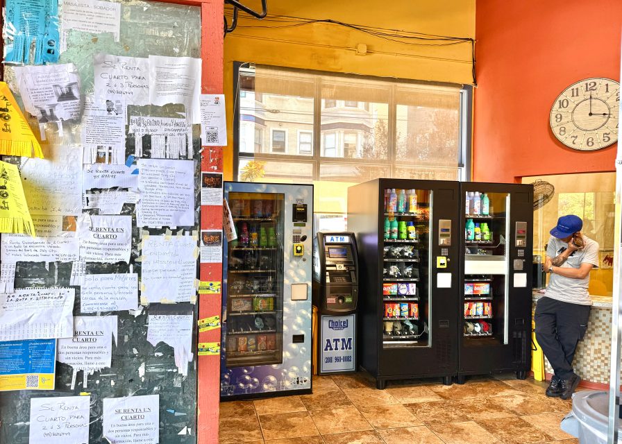 A person stands beside three vending machines and an ATM in a brightly colored room with handwritten notes covering part of the wall. A large clock is mounted above the scene.