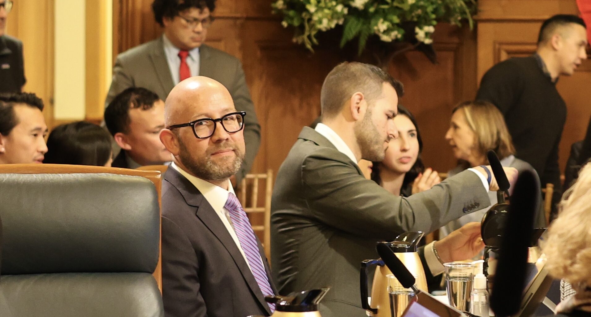 People sitting and conversing in a formal meeting room, with one person in focus wearing glasses and a suit.