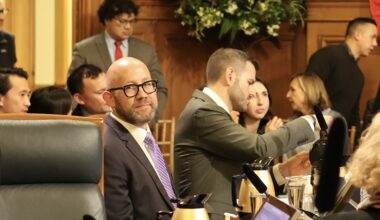People sitting and conversing in a formal meeting room, with one person in focus wearing glasses and a suit.