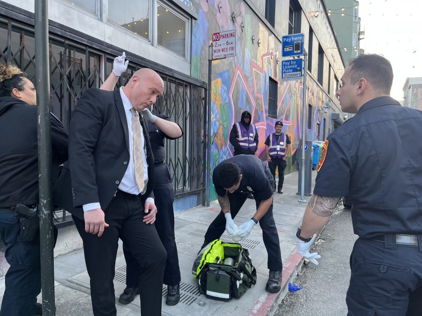 A man in a suit is being attended to by emergency responders on a city sidewalk while others observe nearby.