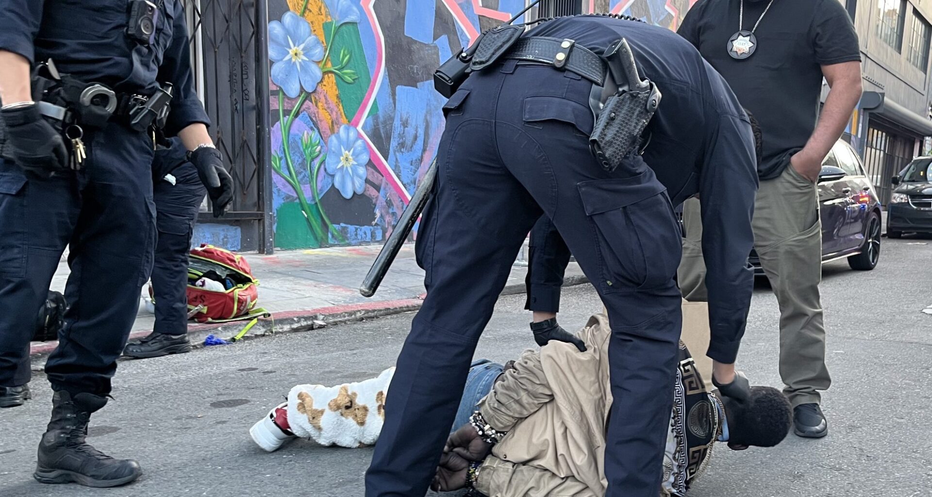 A person is lying face down on a city street while a police officer handcuffs them. Other officers and a plainclothes individual stand nearby. Cars and a mural are visible in the background.