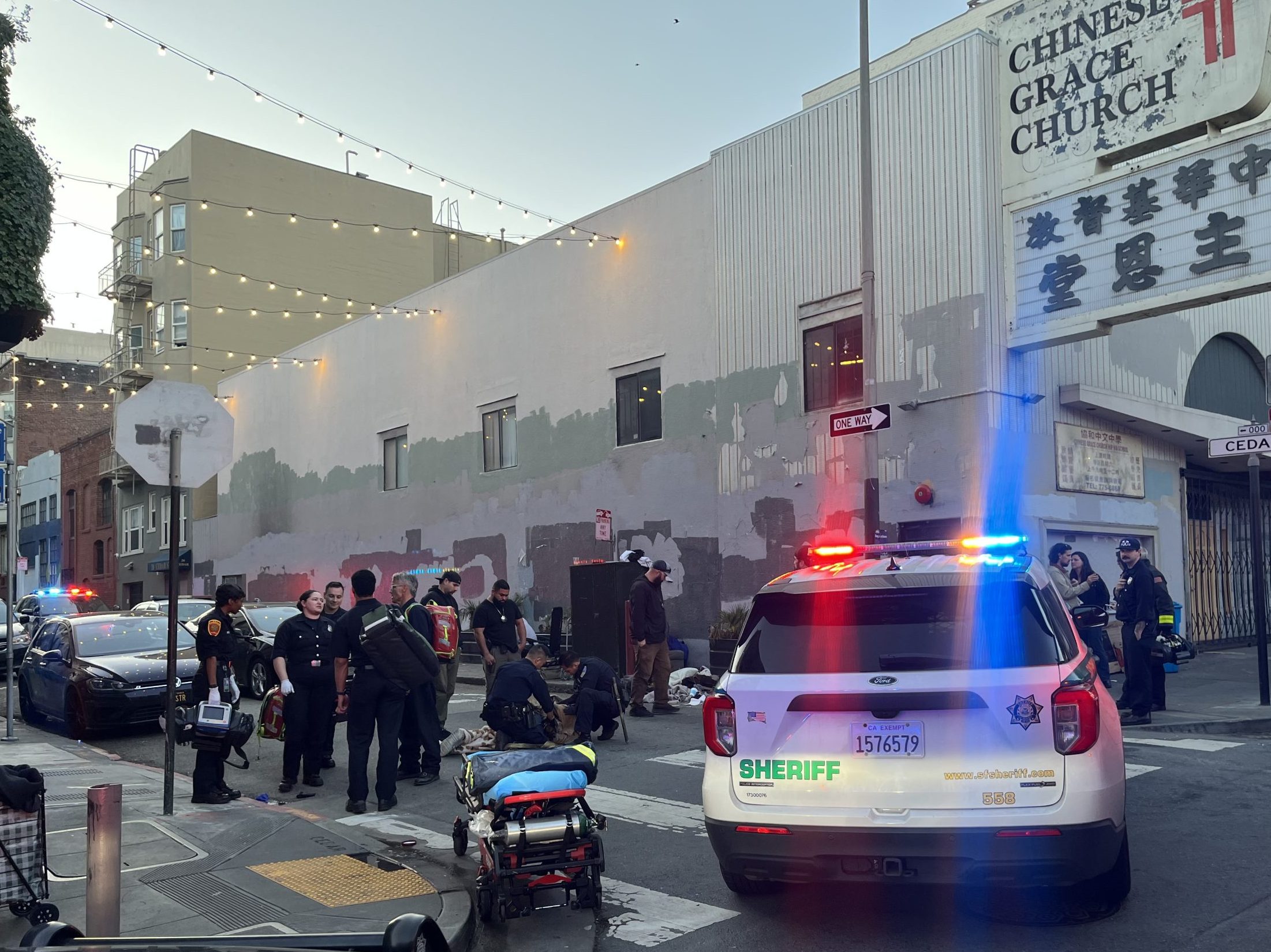 Police officers, sheriff, and paramedics respond to an incident on a city street near the Chinese Grace Church; emergency vehicles have lights flashing.