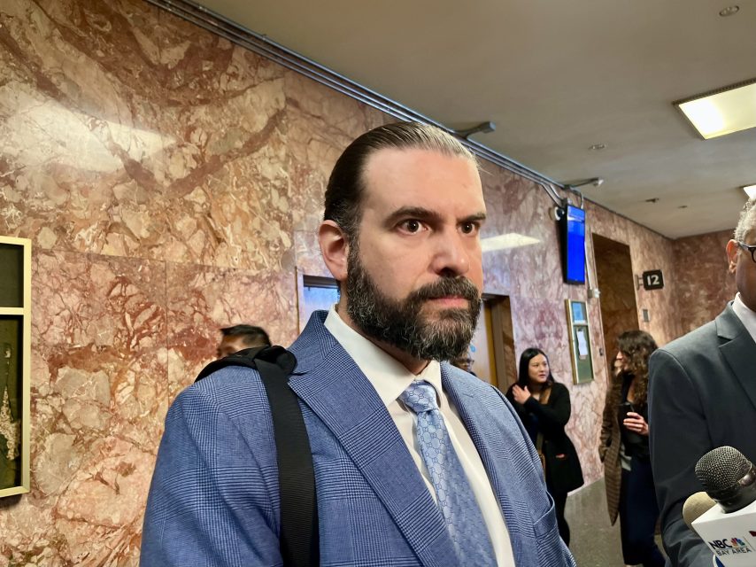 A man in a blue suit and tie stands in a hallway with marble walls, carrying a shoulder bag. Several people talk in the background.
