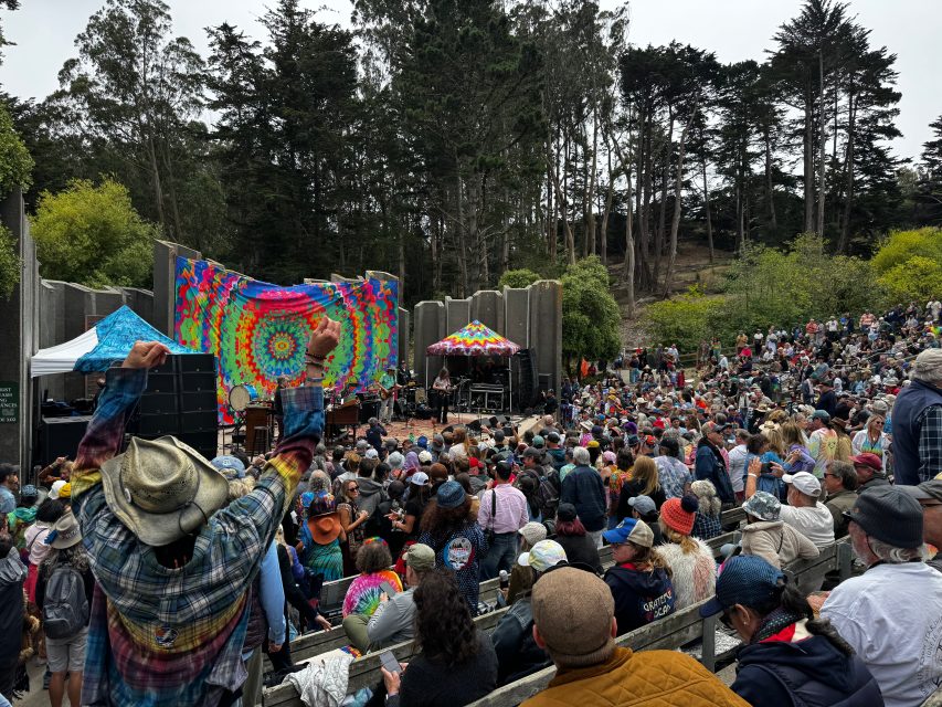 Large outdoor crowd gathers at an amphitheater for a concert with a colorful tie-dye backdrop and surrounding trees.