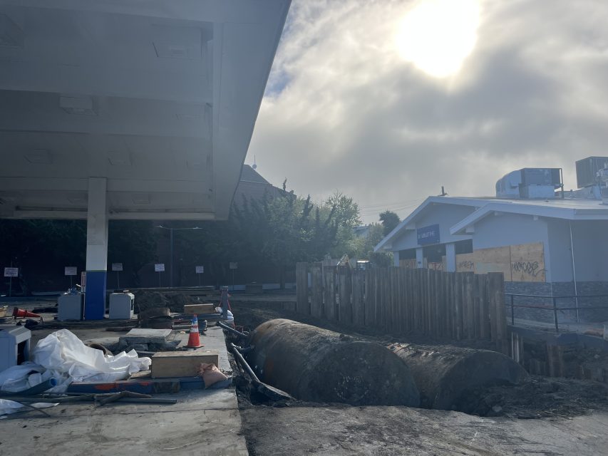 Abandoned gas station under cloudy sky, with construction materials, debris, and large rusted tanks partially buried near a wooden barricade.