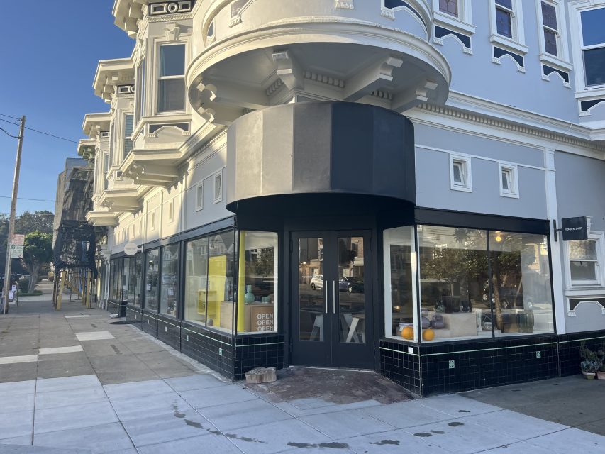 A corner storefront with large windows, black trim, and an "open" sign in the door; shelves with various items are visible inside.