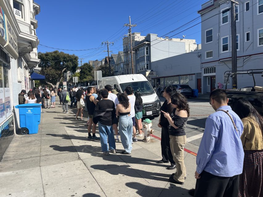 People stand in a long line on a sunny sidewalk in an urban neighborhood, waiting near a row of buildings and a white van.