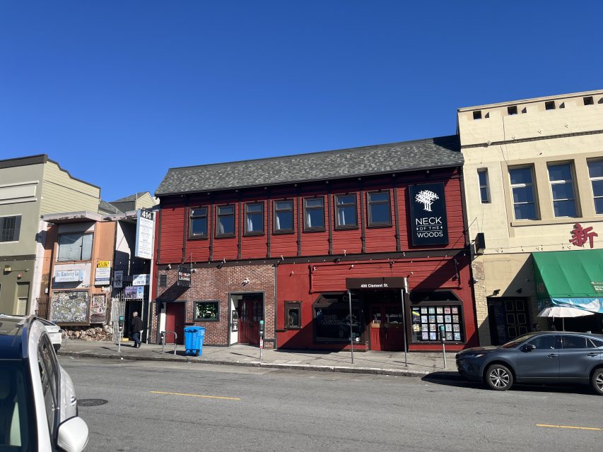A red two-story building houses Neck of the Woods music venue, located on a city street with other storefronts and parked cars under a clear blue sky.