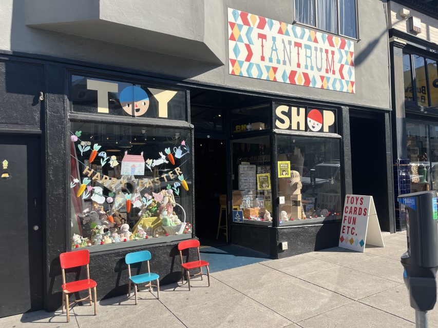 Street view of a toy shop called Tantrum, featuring colorful chairs outside, festive Easter decorations, and a sandwich board listing toys, cards, and other items for sale.