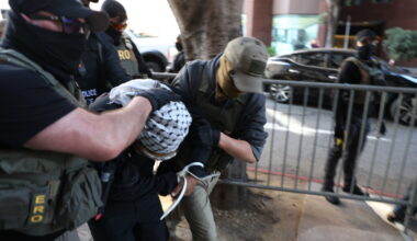 Several masked law enforcement officers detain and handcuff a person in front of a metal barricade on a city sidewalk.