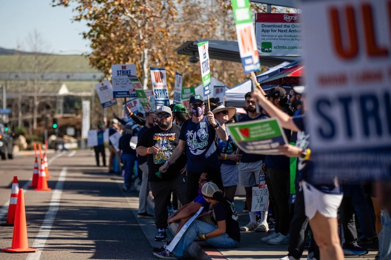 A group of people holding strike signs stand together on a sidewalk beside an empty street with traffic cones.