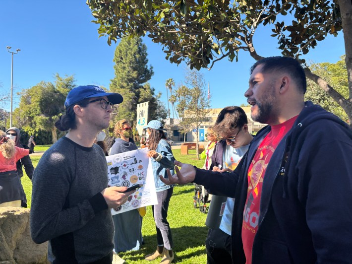 Organizer Kris Rehl and Councilmember Soto-Martinez stand face to face in a park. People in the background hold signs.