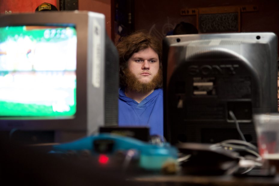 A person with long hair and a beard sits behind two old CRT monitors, focusing intently. The setting appears to be a gaming or computer event.
