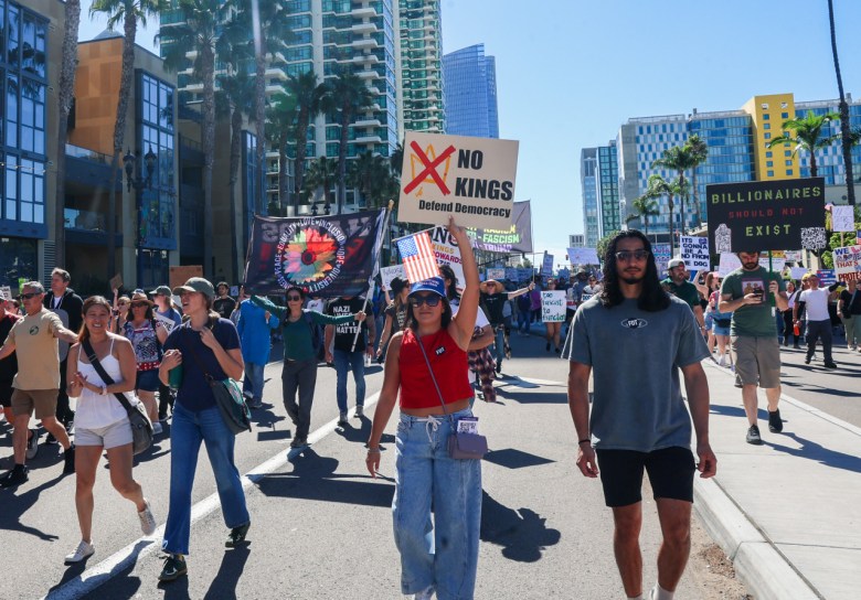 Marchers walking on a downtown street bearing "No Kings" signs and another that reads "Billionaires should not exist."