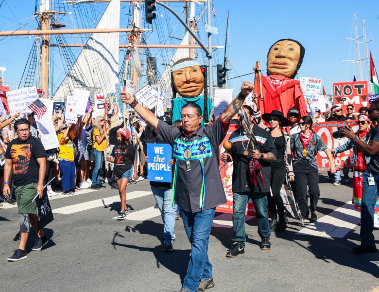 With the tall ships on the bayfront seen in the background, Native American marchers hold puppets while standing among dozens of people with signs and a few with U.S. flags.