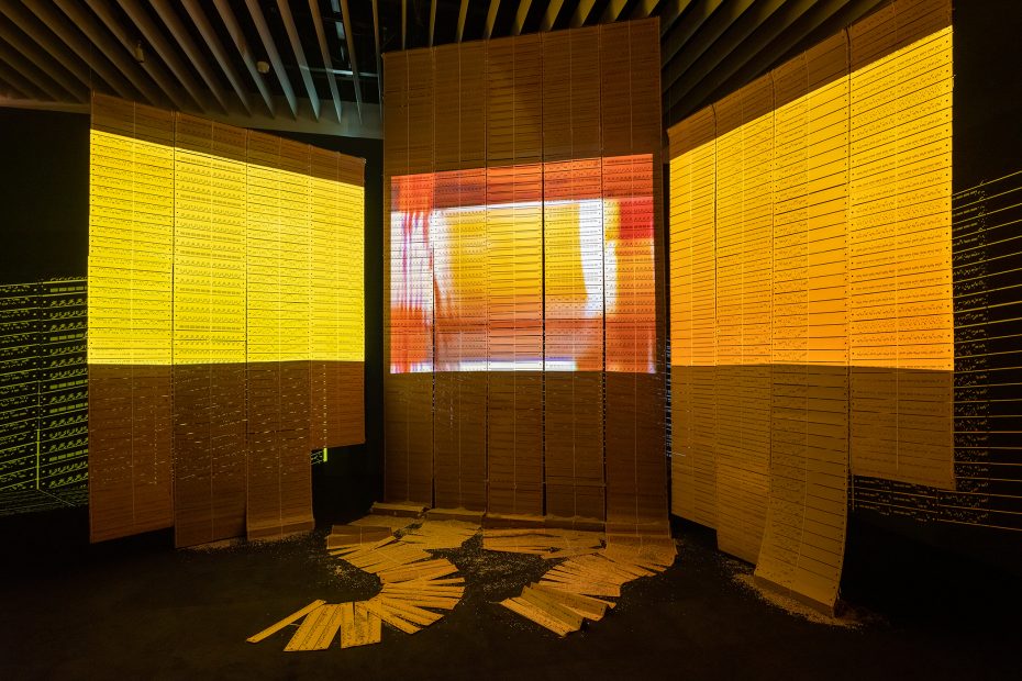 Large illuminated yellow panels and sheets are arranged vertically and spread on the floor in a dark room with a ceiling of parallel beams.