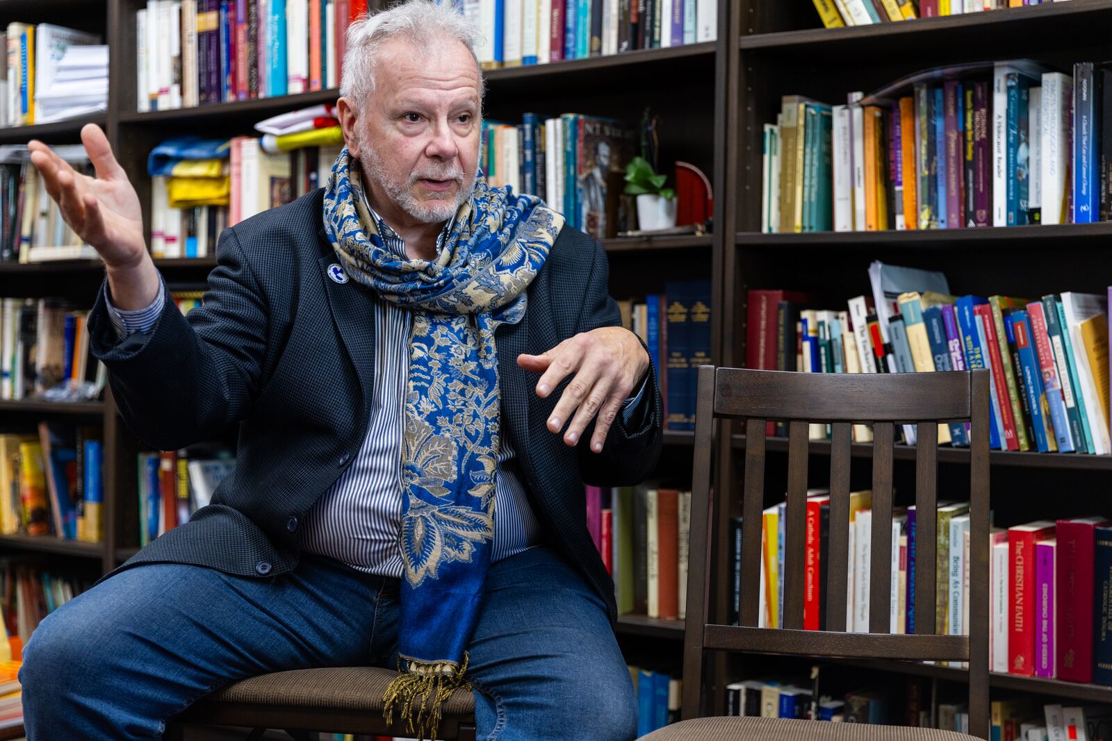 A man wearing a dark suit jacket and a bright blue scarf sits in a chair, waving his hands as he talks, in front of a bookcase.
