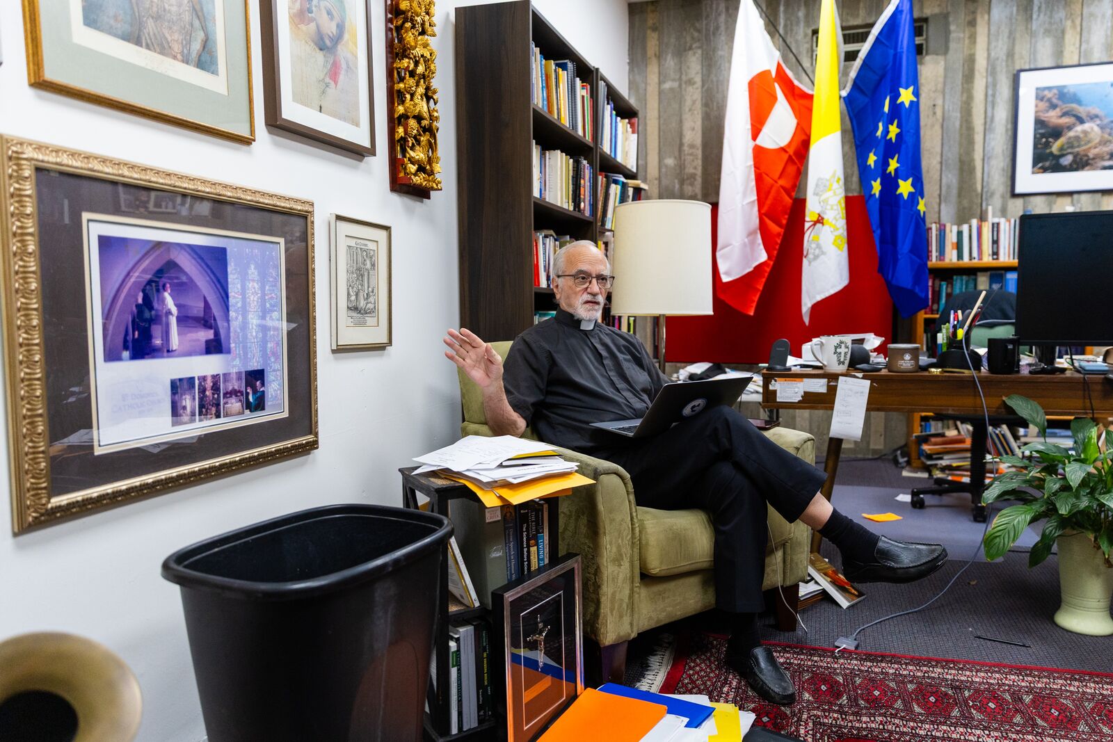 A priest sits in a chair inside an office. Several flags, including the Vatican flag, are visible in the background. A black trash can is visible in the foreground.