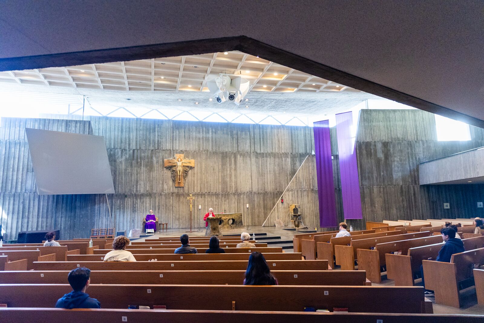 People attend a mass inside a church sanctuary with gray concrete walls. Sunlight pours in through gaps in the wall near the ceiling