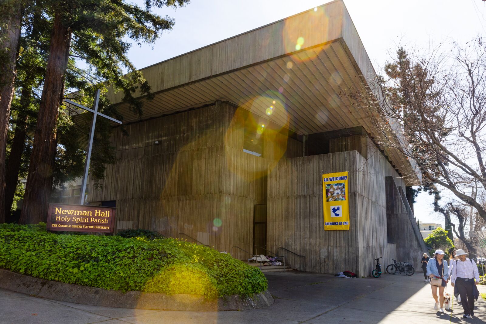 A view of the concrete exterior of a church; people walk past it on the sidewalk