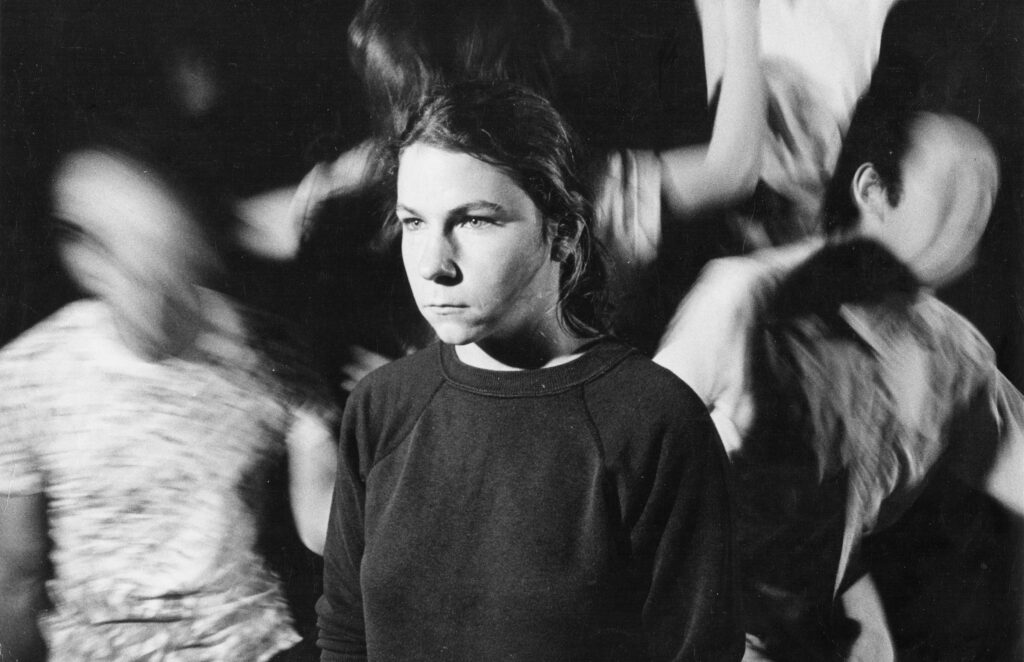 A black-and-white theatrical photo of Nina Ruscio at UC Berkeley in the early 1980s. She stands still and somber in a dark sweatshirt, surrounded by several figures in motion blur that create a sense of chaotic energy.