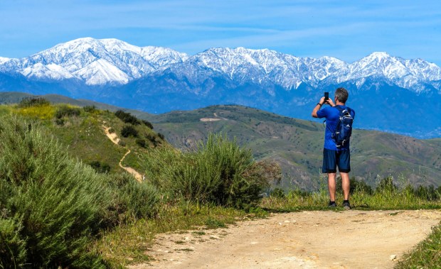 A hiker stops to take a photo of the snow-covered San Gabriel Mountains while hiking along Old Weir Canyon Trail in Anaheim Hills, on Sunday afternoon, February 22, 2026. (Photo by Mark Rightmire, For the Orange County Register/SCNG)