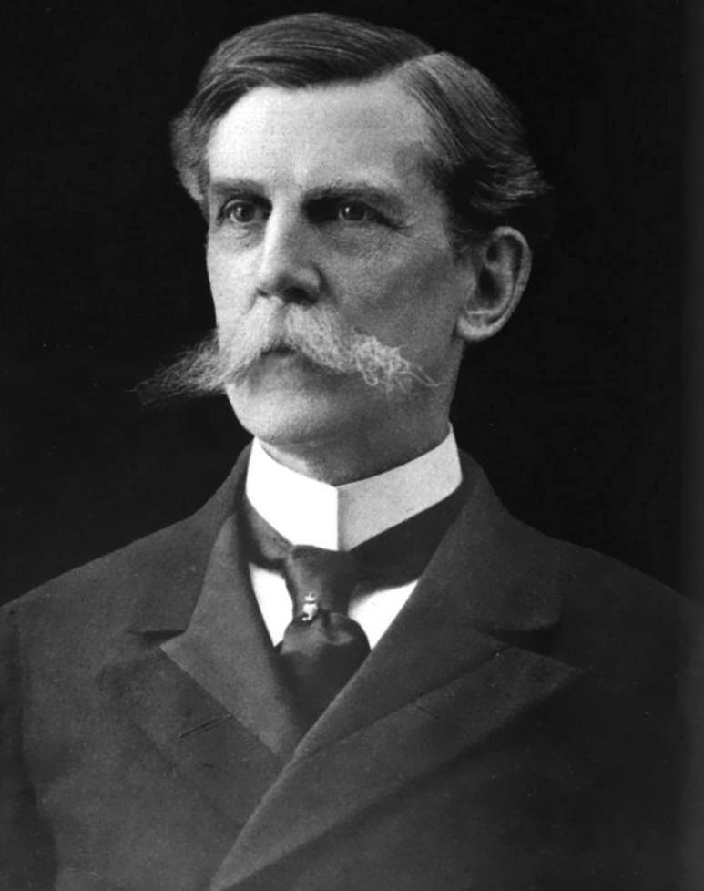 A black and white headshot of Oliver Wendell Holmes in 1902, with a long handlebar mustache, wearing a dark suit jacket over his white shirt and dark tie.
