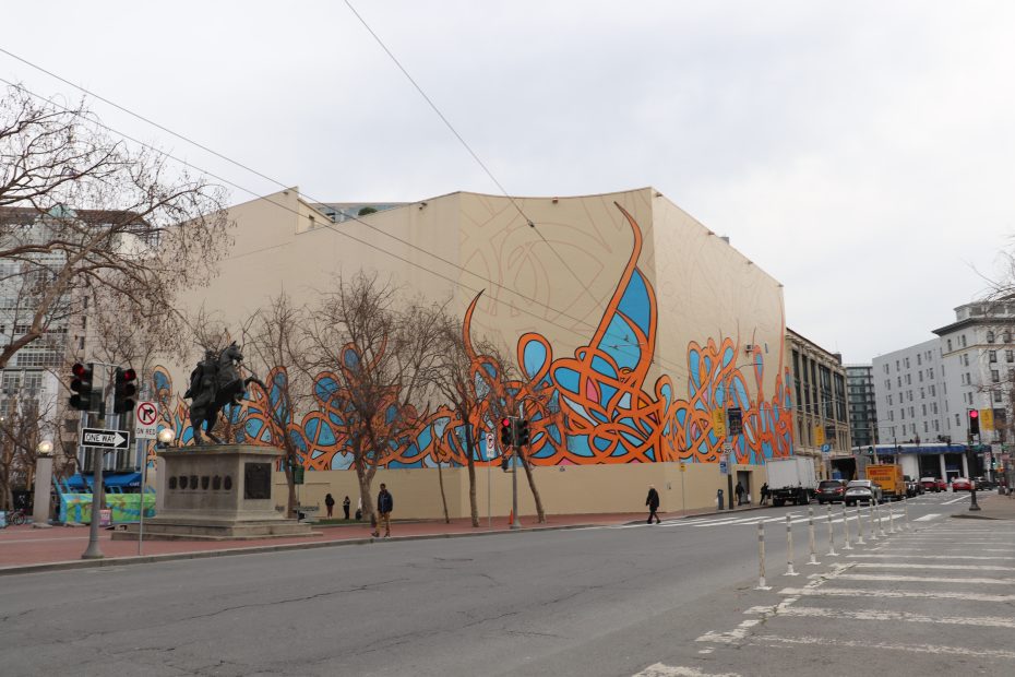 A large beige building features an abstract blue and orange mural; leafless trees and a statue stand nearby, with pedestrians and cars on the street.