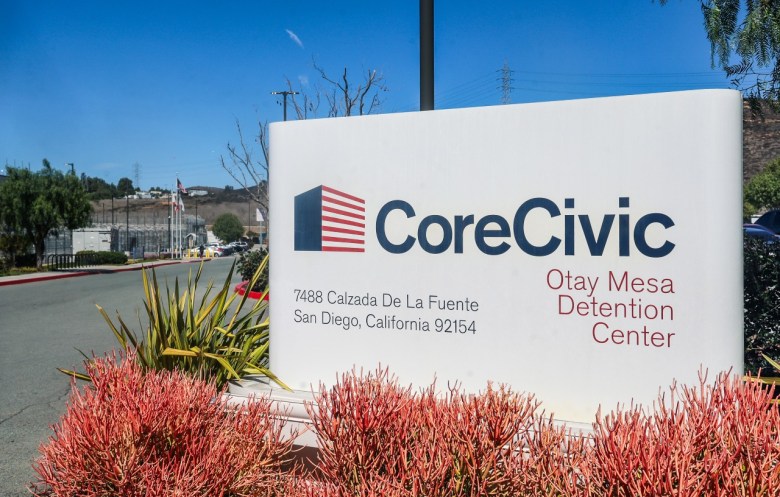 Sign for "CoreCivic Otay Mesa Detention Center" with address, surrounded by plants and flags in the background.