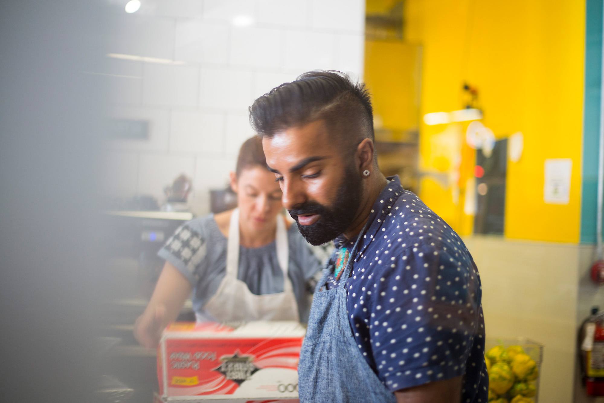A man in a blue apron looking down in front of a restaurant kitchen, where a woman is prepping food.