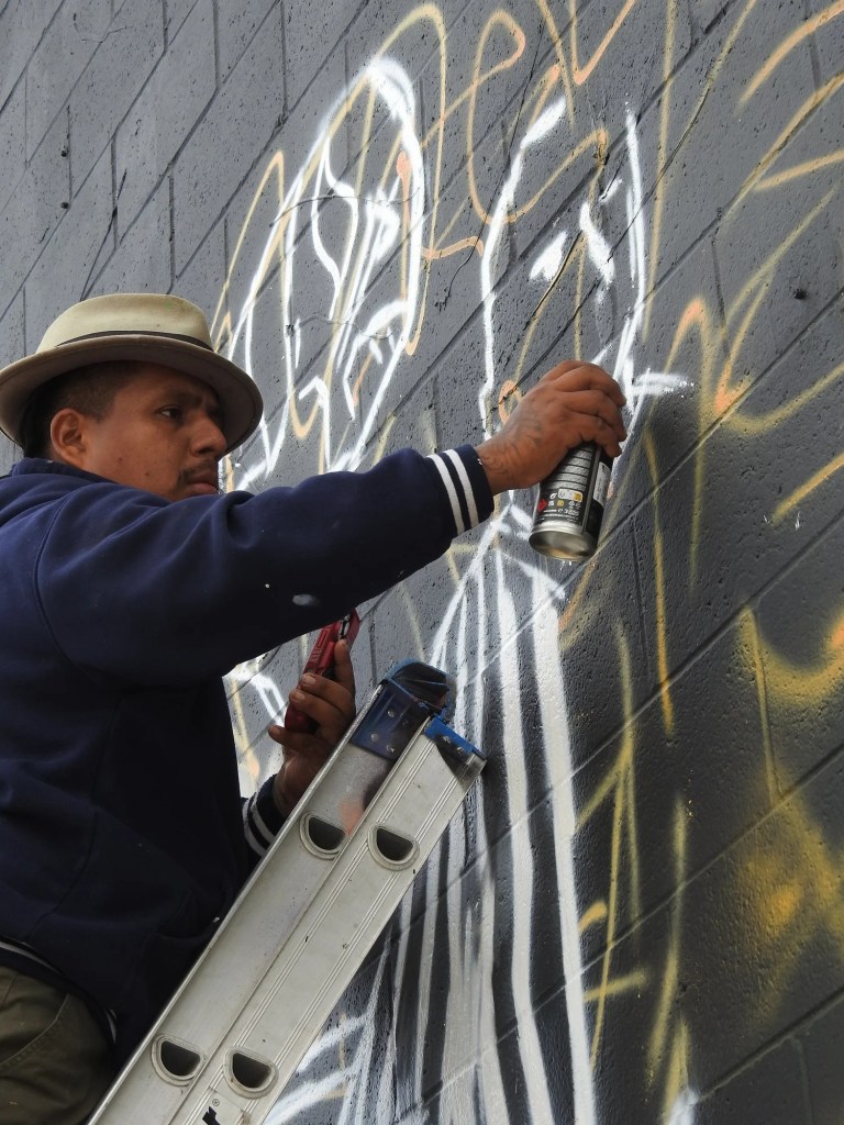 Person on a ladder spray painting abstract art on a dark brick wall.