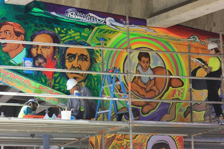 A vibrant mural with human figures and abstract designs on a wall, with people working on scaffolding in front.